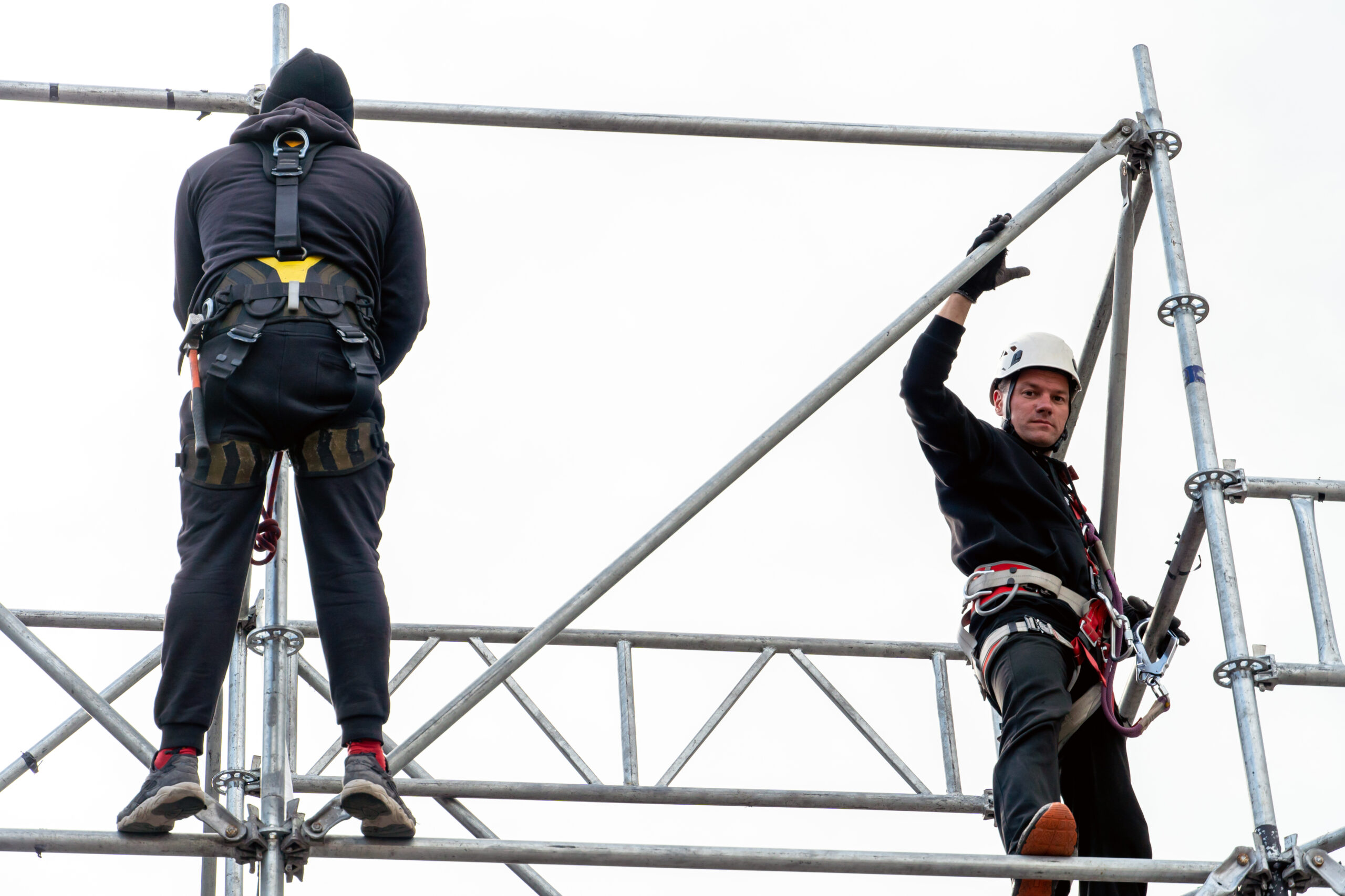 An industrial climber wearing a helmet and a protective belt is tied with a rope to a support at a high altitude. A specialist assembles a stage structure from a modular system of metal scaffolding.